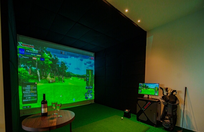 ゴルフシミュレーター。大きなスクリーンに映し出されたゴルフコースと、手前にはゴルフバッグや飲み物が置かれたテーブルがある。